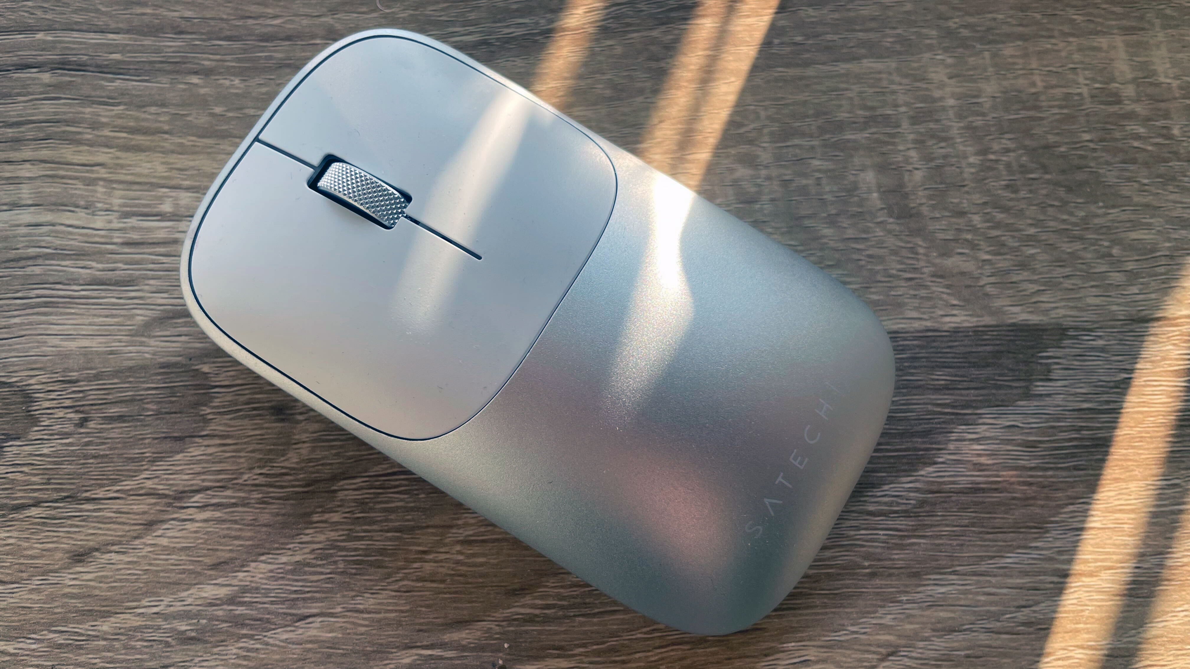 A silver grey Satechi Slim EX3 keyboard and Slim EX mouse on a wooden desk with sunlight shining on it from a window in the background