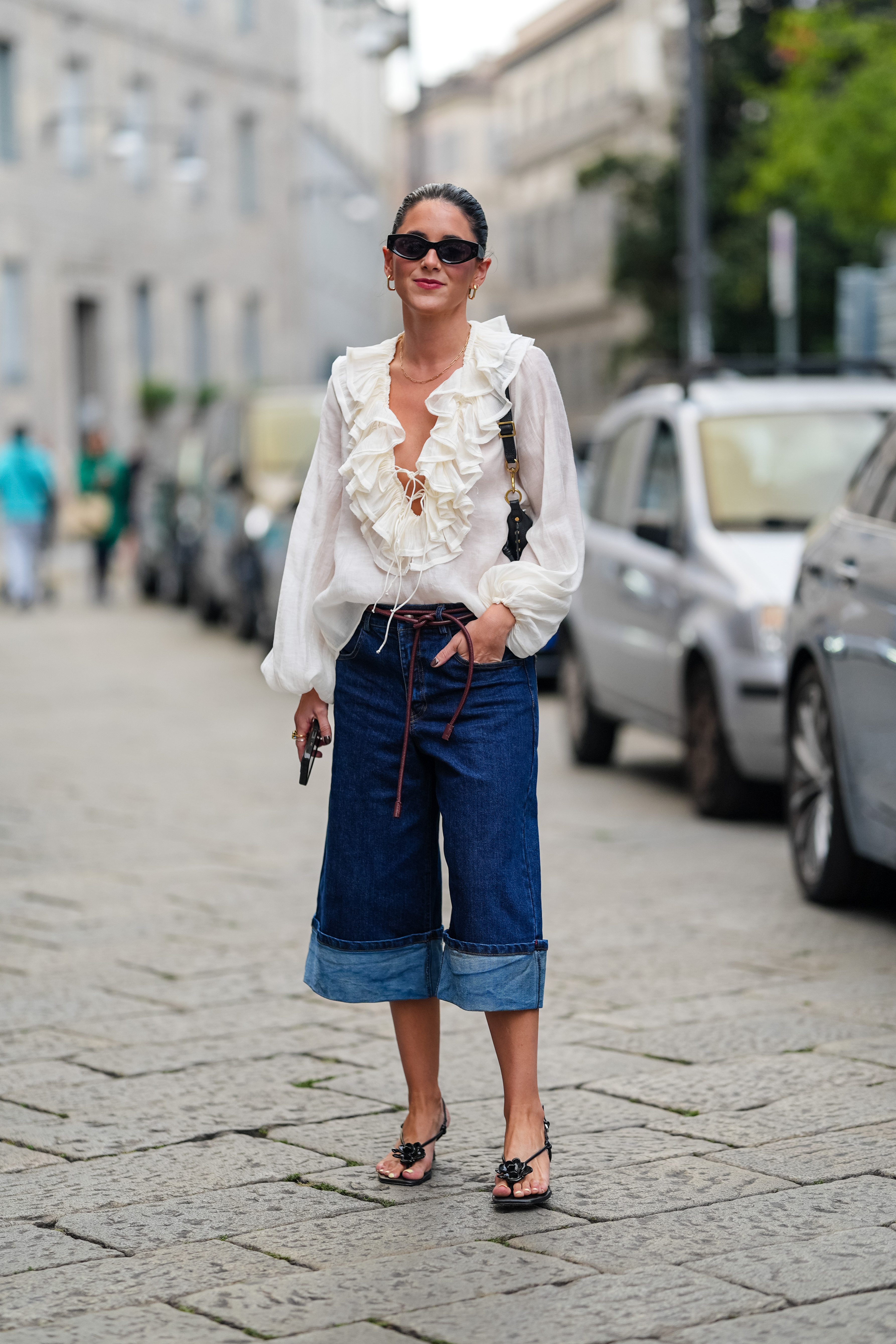 A guest wears black sunglasses, gold earrings, white ruffled long sleeve shirt, black leather bag, dark navy blue denim jean folded pants, black flip flop heels leather shoes with floral detail, outside Sportmax, during the Milan Fashion Week Spring/Summer 2024-2025 on September 20, 2024 in Milan, Italy