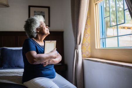 A widow looks out a window, holding a portrait of her spouse.