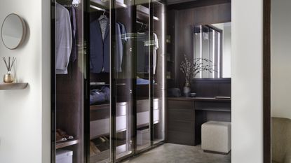 A walk-in closet with dark wood, organized clothes, and a built-in dresser with a beige, square-shaped stool