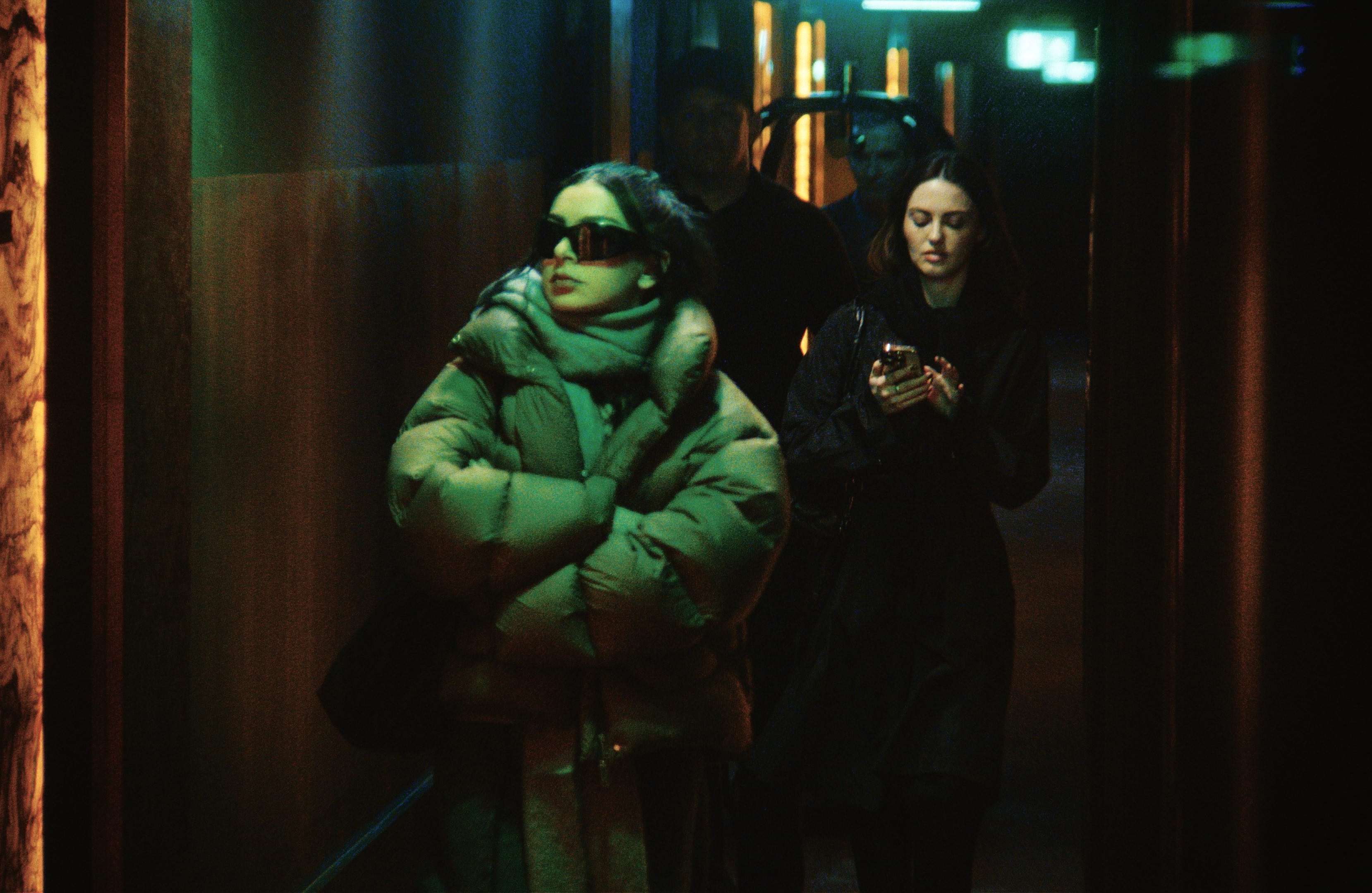 charli xcx wears a puffer coat and sunglasses as she walks down a dark hallway and an assistant follows behind her in a still from the movie the moment