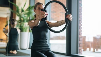 woman wearing black vest and leggings holding a black pilates ring out in front of her towards the camera. there's a mirror behind her and large window to the side. 