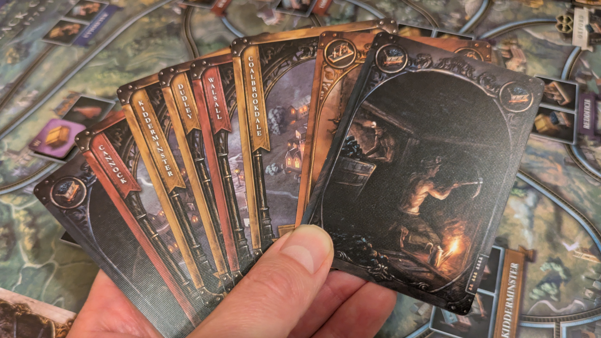 A hand holds out a spread of Brass: Birmingham cards, with the map board visible in the background