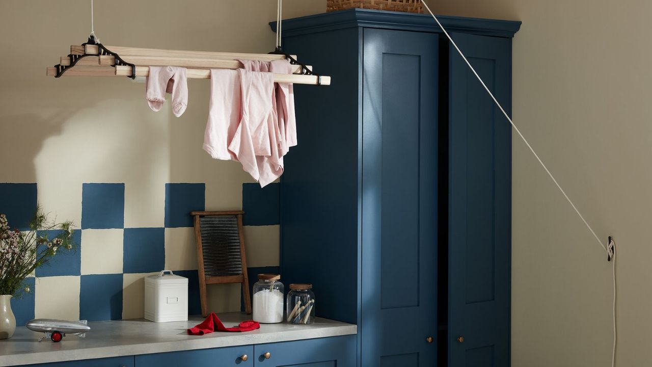 Dark blue painted cabinets with matching blue and white tiles in a small laundry room. A ceiling-suspended drying rack above, with some pink clothes on it. 