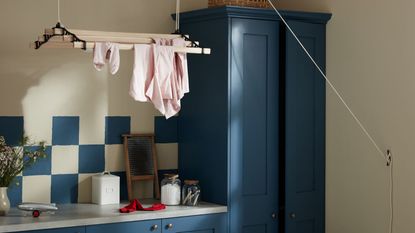 Dark blue painted cabinets with matching blue and white tiles in a small laundry room. A ceiling-suspended drying rack above, with some pink clothes on it.