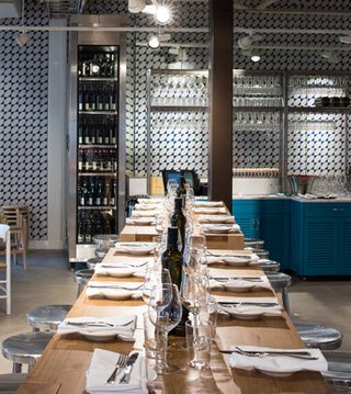 Hornhuset, Stockholm, Sweden. A restaurant with a row of decorated tables with round silver chairs, a wine fridge and wall shelving.