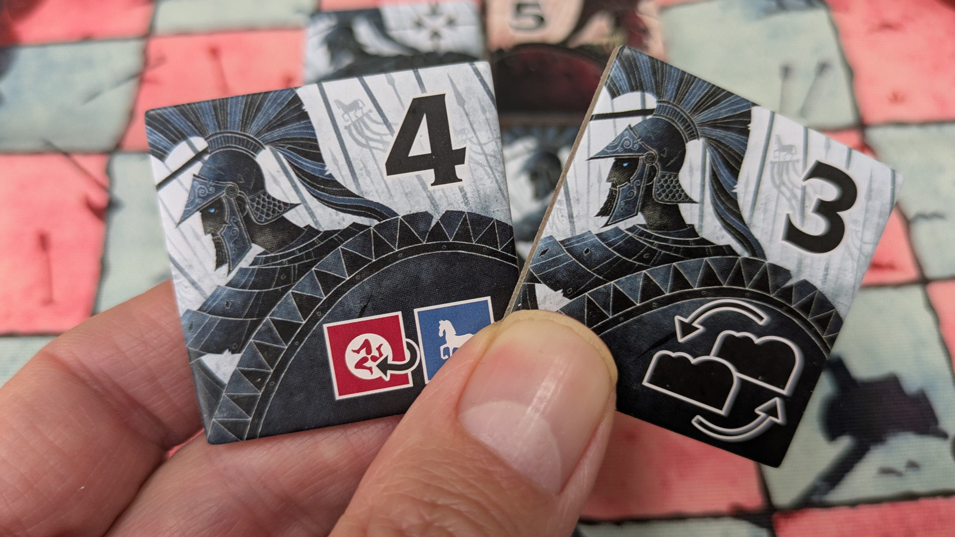 A hand holds two numbered tiles with a Greek warrior on them