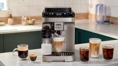 A De'Longhi coffee machine in black and stainless steel with a glass of iced coffee on the drip tray beside an automatic milk dispenser. 