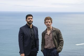 A posed shot of Barney Crozier (Robert Gilbert) and Jim Bergerac (Damien Molony) looking directly at the camera, with the sea in the background. Barney is wearing a suit with no tie and has his left hand in his pocket. Jim is wearing an open-necked shirt and suit trousers, with a unmatching jacket over the top, and has both hands in his pockets.