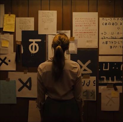 a woman with her back to the camera stands in front of a wall of papers and cryptic drawings