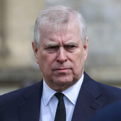 Prince Andrew wears a black suit with a white shirt and black tie and has white gray hair and is scowling