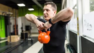 A man in a black t-shirt performs a kettlebell upright row in a gym. He's standing, with both hands on the kettlebell, and a fitness tracker on his wrist. His elbows are bent and held up slightly higher than his shoulders, so the kettlebell rests at chest height.