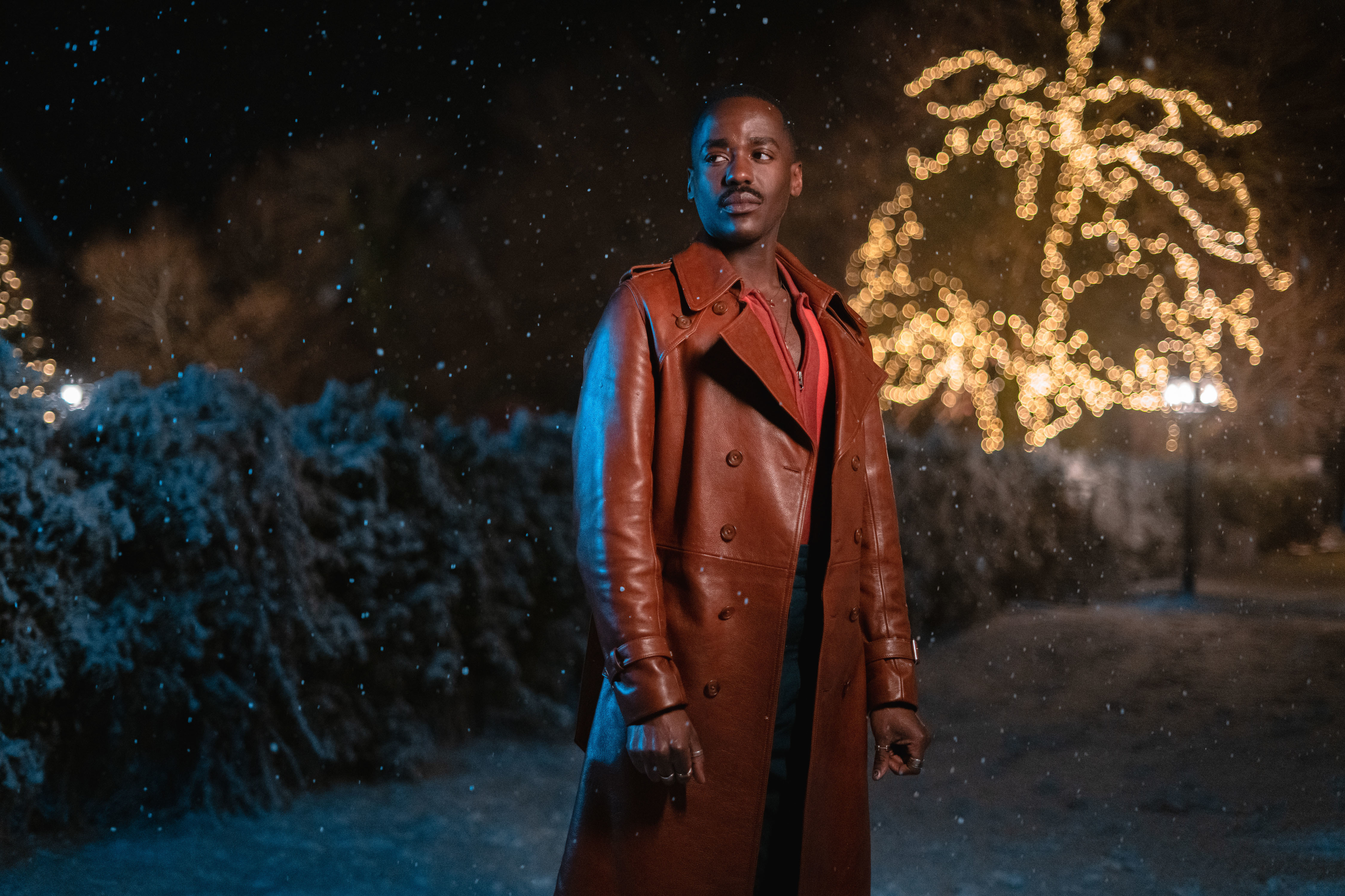 The Doctor (Ncuti Gatwa) stands in a garden with snow falling all around him and Christmas lights in a tree. behind him. He is wearing a long brown leather coat.