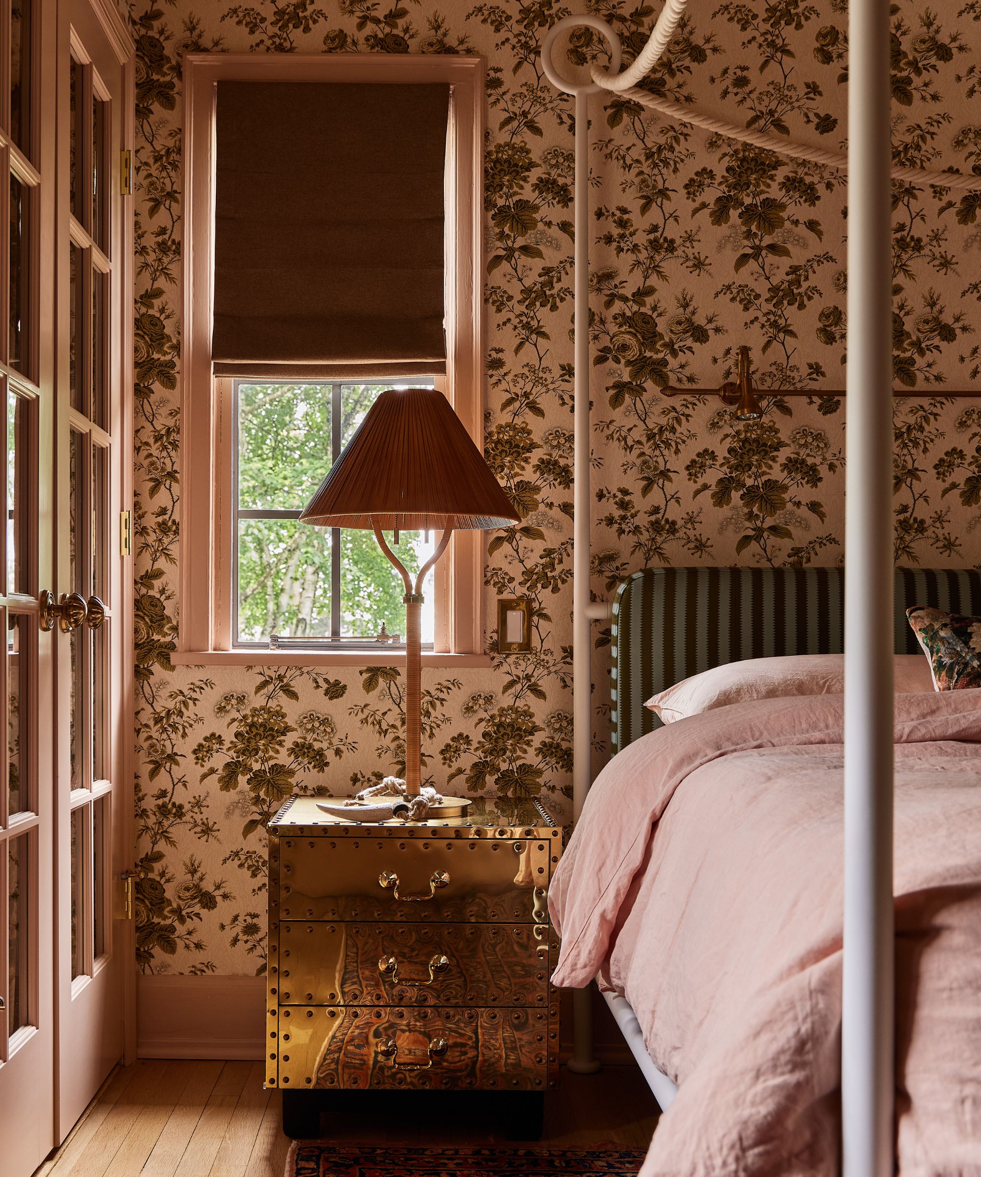 a floral wallpapered bedroom with gold mirrored nightstands with a wicker table lamp sat in front of a small window. The bed is a canopy style with stripe headboard and rope detailing