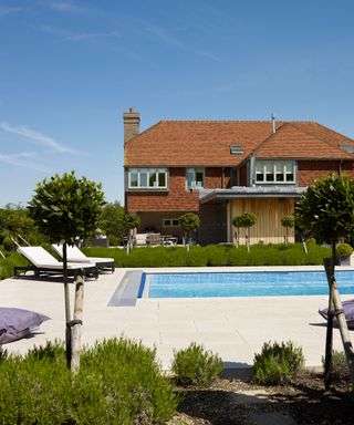 A swimming pool with a paved surround, white loungers and border planting in front of a house