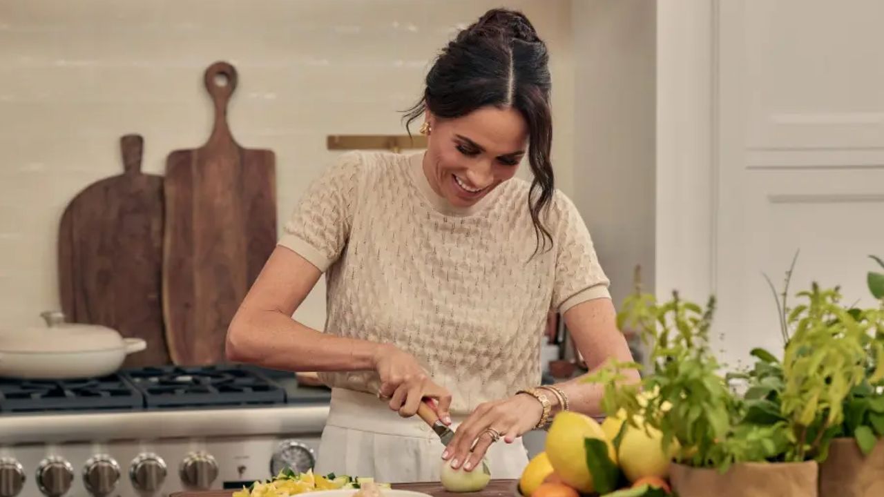 Meghan Markle wearing a beige short sleeved sweater cutting lemons on a table in a white kitchen