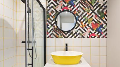 Small bathroom with white tiled walls, aviary geometric wallpaper, yellow grout, yellow sink on white cabinet and black metal hardware including on the the shower and screen, mirror and faucet
