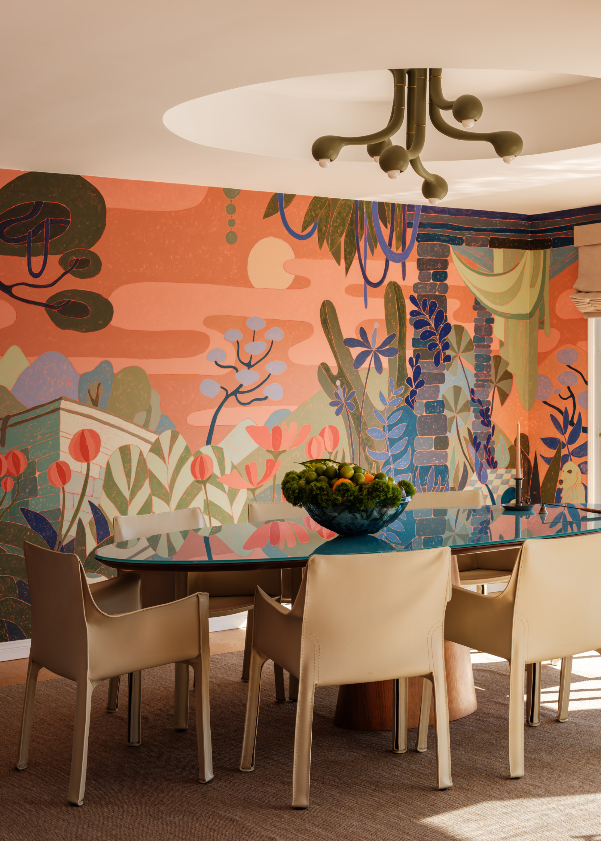 A bright living room with a green overhead light fixture, a glass table, a set of cream chairs, a bowl of fruit and vegetables, and a colorful mural accent wall
