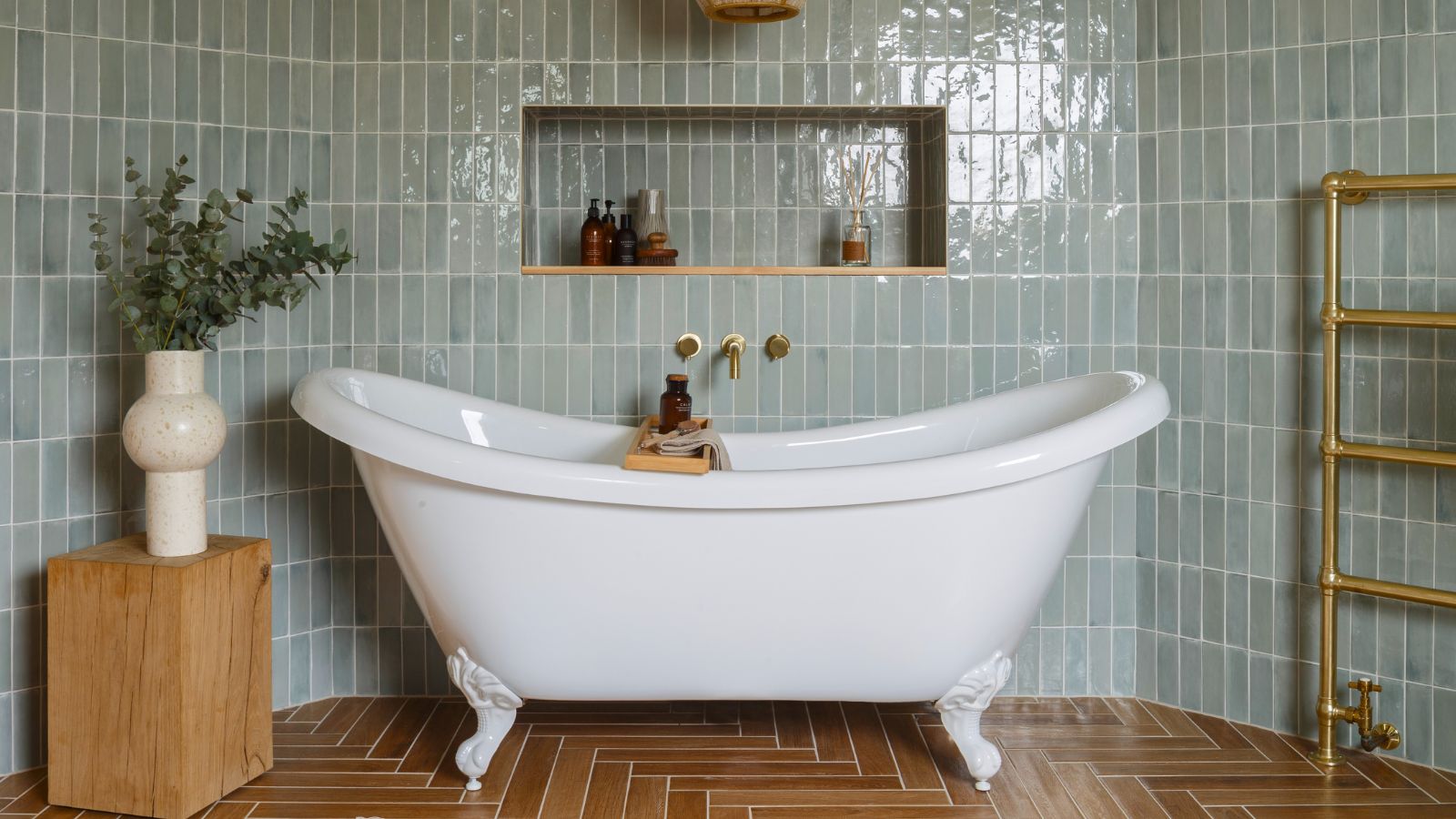 A classic white roll top bath tub in a large bathroom with blue tiled walls and a red herringbone tiled floor. 