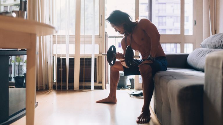 Muscular man curling a dumbbell while sitting on the sofa in a living room