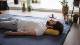 A man lies on a yoga mat with a pillow under his head. His eyes are closed and candles are lit behind him. We can also see a large window and plants in the background.