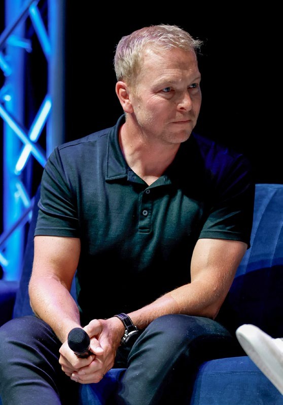 Chris Hoy seated, wearing a dark polo shirt, microphone in hand