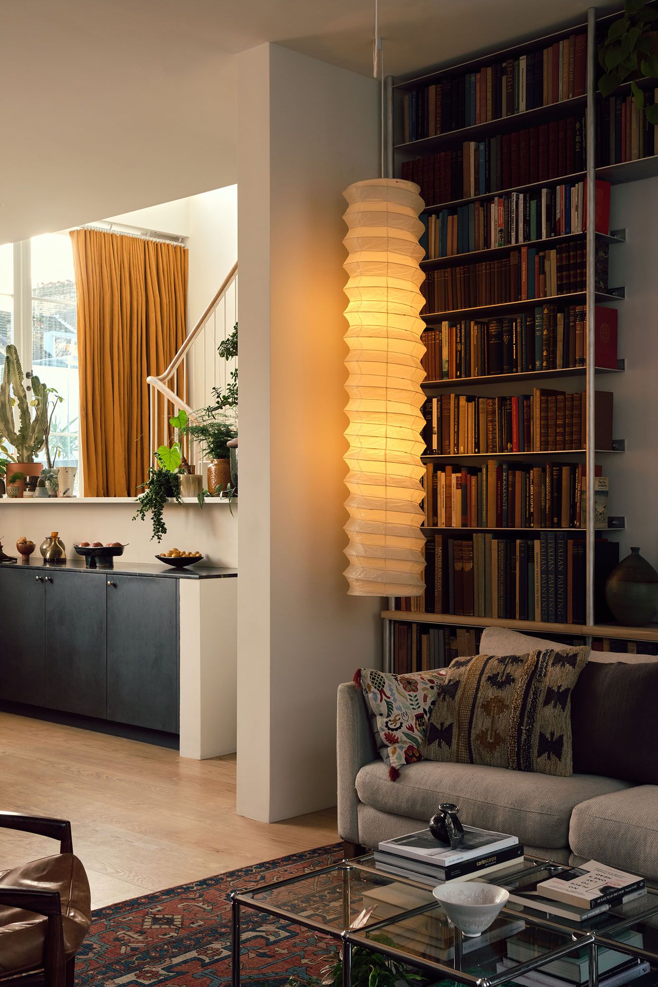 Interiors of a beautifully designed, spacious home with wooden floors, neutral decor, and lots of colorful accessories and furniture, including thriving plants and a golden-glowing paper lamp.