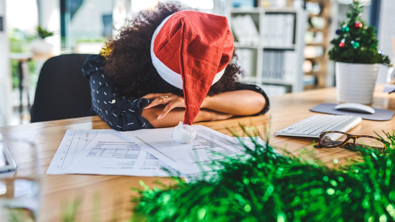 A woman wearing a Santa hat sits with her head down on her desk.