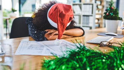 A woman wearing a Santa hat sits with her head down on her desk.