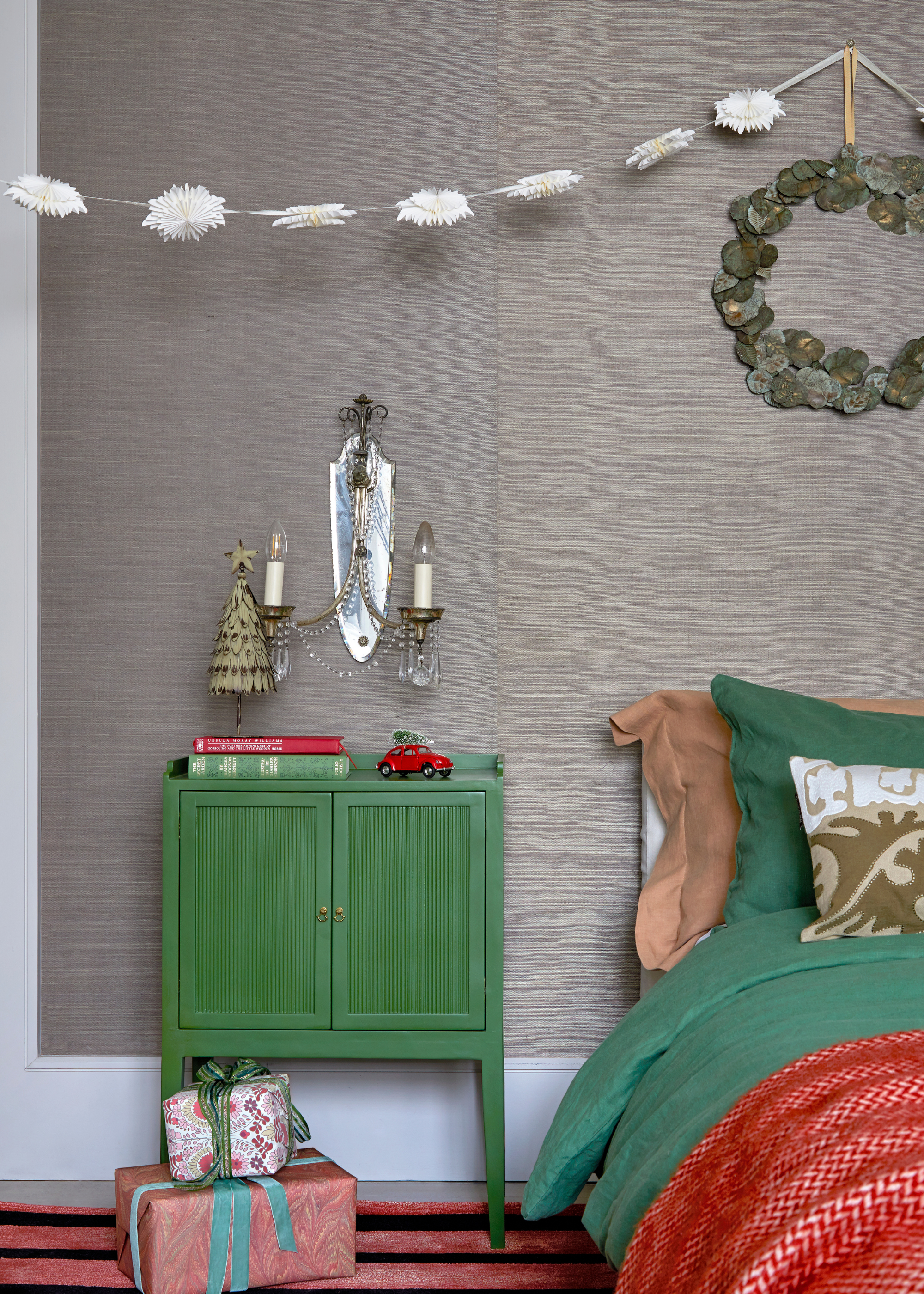 A bedroom decorated for Christmas with red and green accents, a wreath above the bed and a white feather garland, with presents by the nightstand and a christmas tree ornament on top of the nightstand