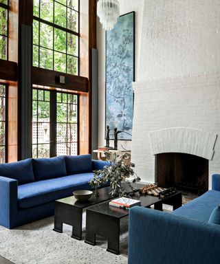 A white living room with double height ceilings and floor-to-ceiling windows framed with wood. A blue sofa and glass coffee table