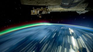 the curvature of earth rises upward just over halfway up the image, lights streaking across its surface like a fast moving train. Blue and red layered aurora are seen over the left half of the curve. A space station module hangs from above, medal and full of copmonents. Streaked stars fill the black void between the vessel and planet.