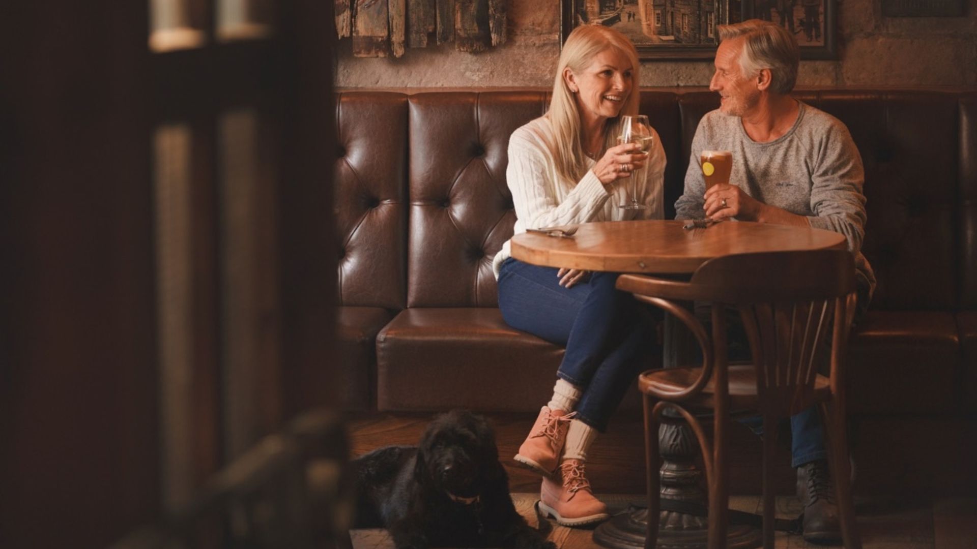 A mature couple enjoy a drink together in a cosy, traditional pub while their dog sits beside them on the floor.