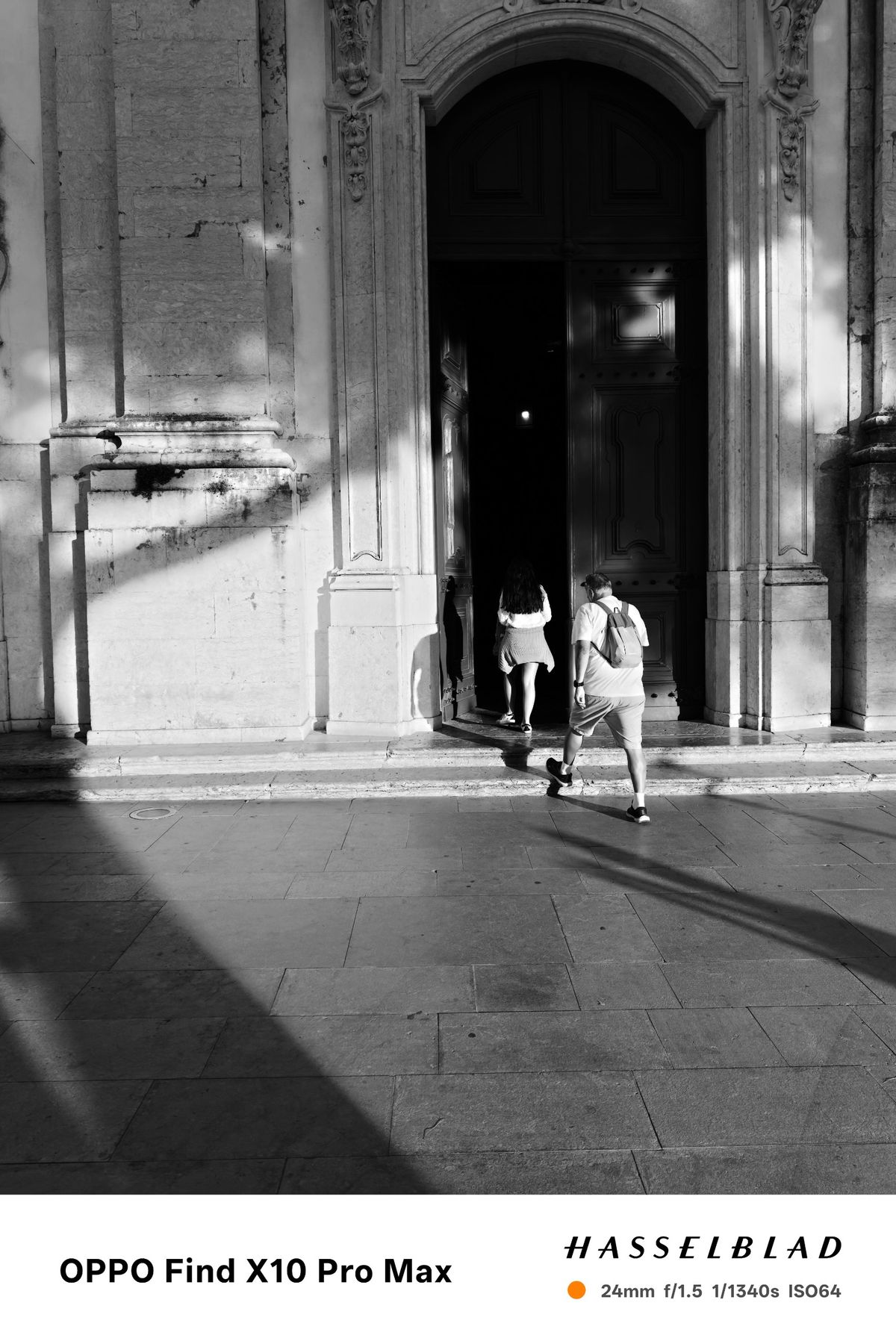 Black-and-white view of two visitors entering a large arched doorway with long shadows across the pavement.
