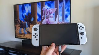 Switch OLED plugged into AceFast cable with game showing up on TV behind it.