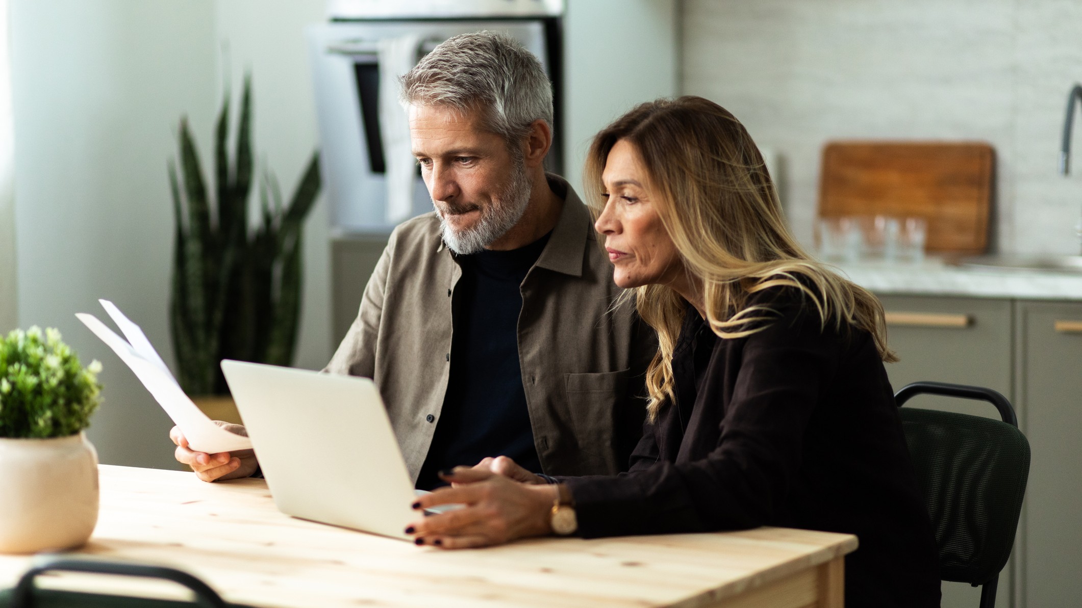 An older couple discusses finances or other serious matter in front of an open laptop on their kitchen table. The image has been modified for 16:9 aspect ratio.