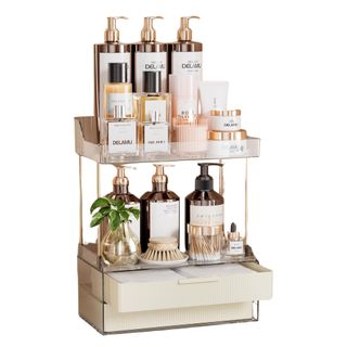 A two-tier bathroom organizer with drawers and rose gold metal accents, filled with glass bottles with rose gold lids, and scrubbing brushes