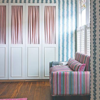 A bedroom covered in a striped wallpaper with a striped sofa and a white wardrobe with pink fabric inserts