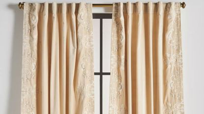 Anthropologie Harlowe curtain panels in Ivory hanging from a decorative brass curtain rod over a tall windowpane.
