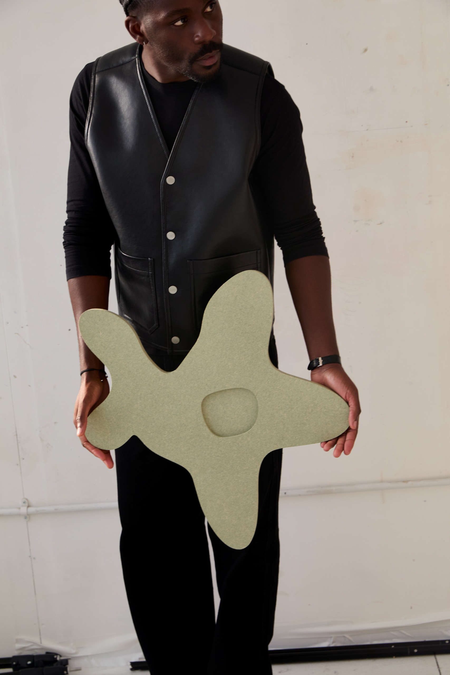 A young man poses in his industrial studio dressed in casual smartwear, sitting or standing in front of a gray wall with his sculptural creations.
