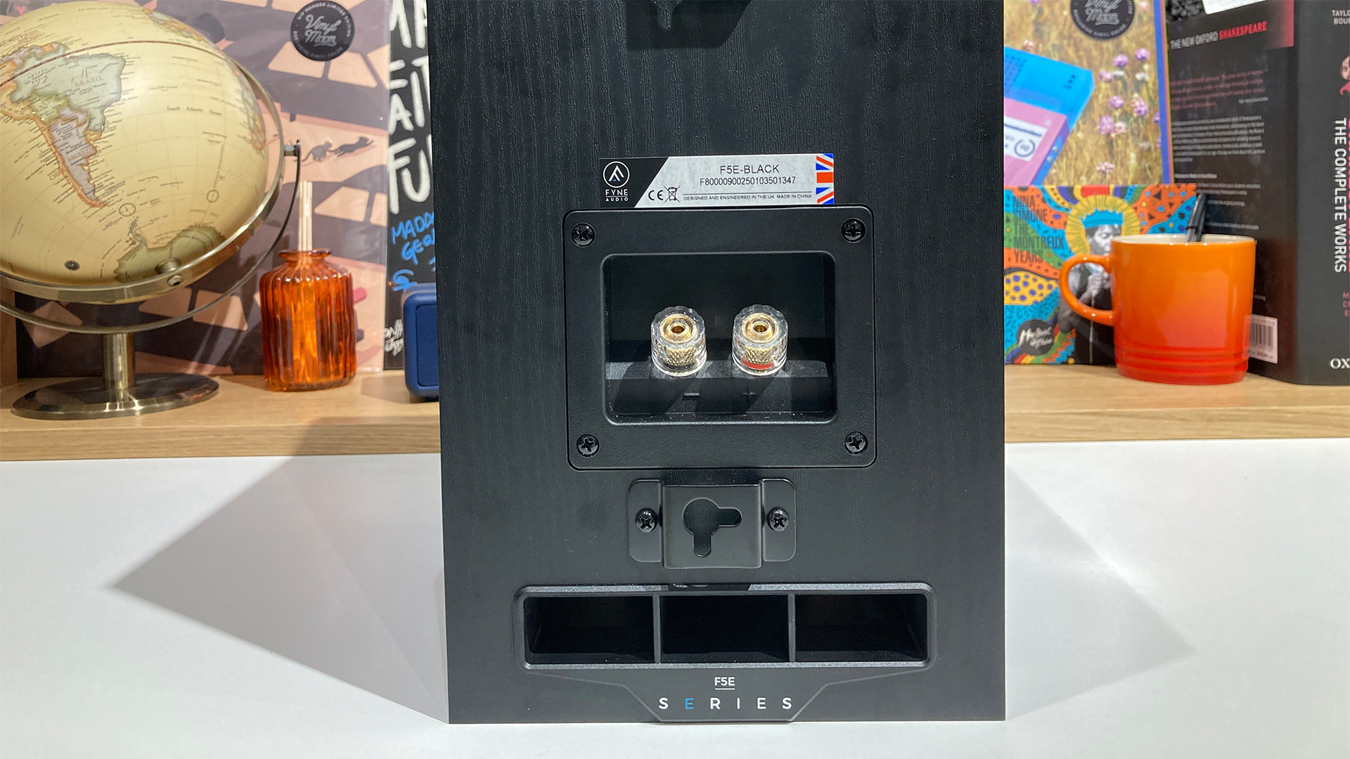 Fyne Audio F5E standmount speakers on white table in front of busy bookcase, rear of speakers showing binding posts