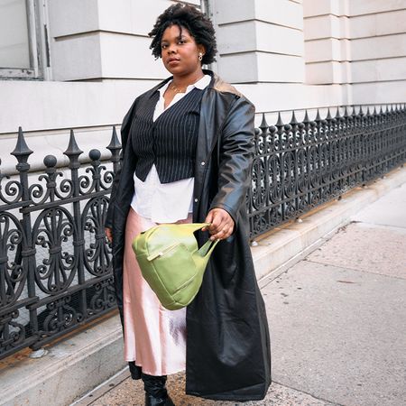 Chichi wears a layered loo consisting of a satin dress, button down shirt, vest, and faux leather trench, and a green purse