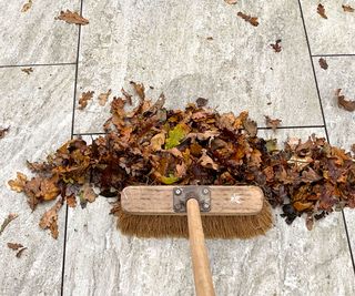 A wooden broom being used to sweep up leaves on a light coloured patio