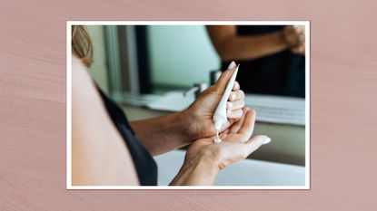 Image of woman squeezing cream out of white makeup tube into the palm of her hand, whilst standing in front of a mirror, on a dusky pink background