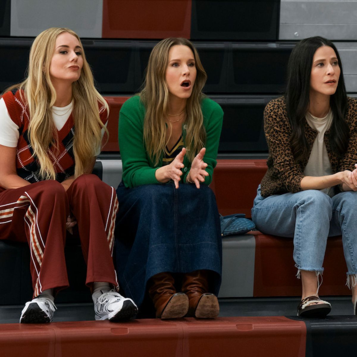 Kristen Bell and co-stars sitting on the bleachers. She's wearing a a green cardigan, a denim skirt and brown boots.