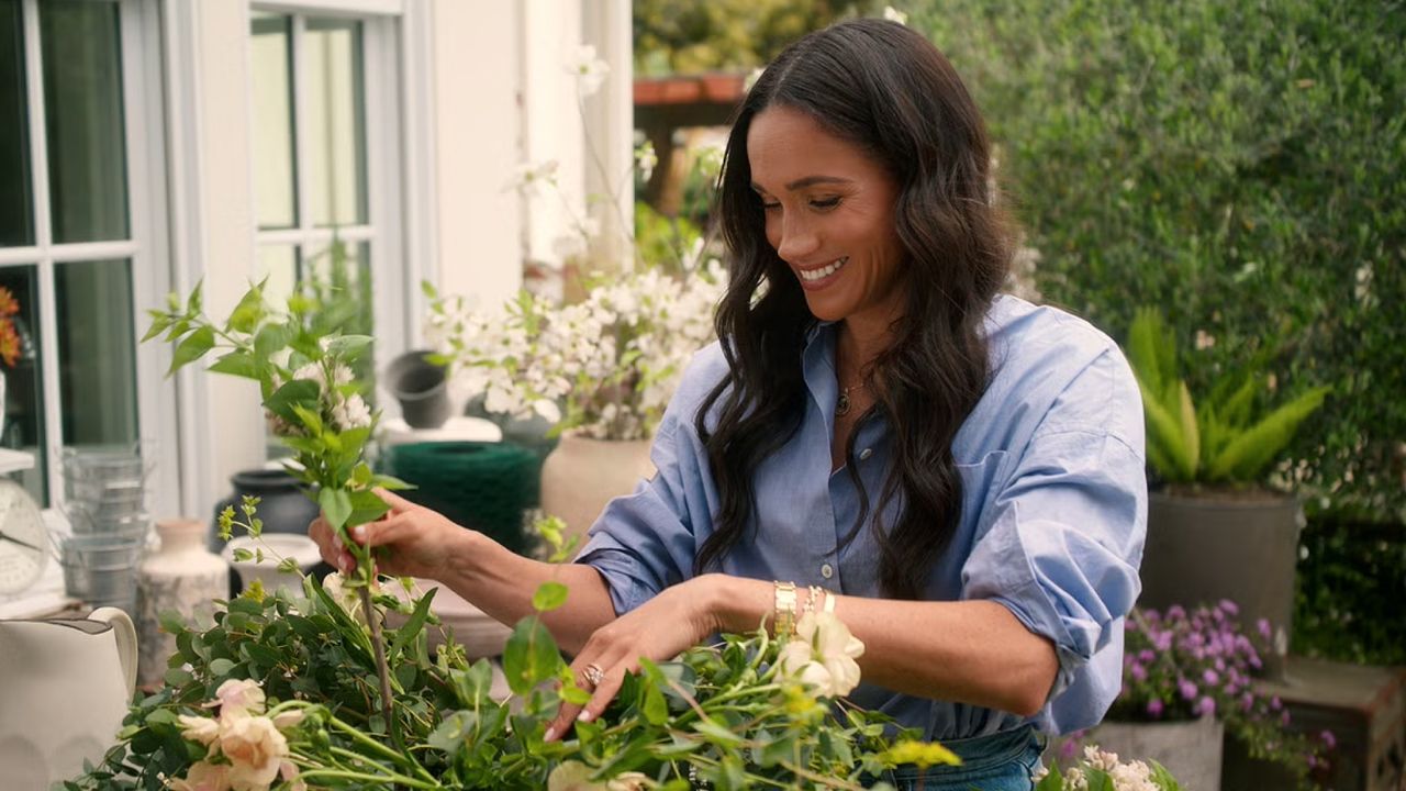 Meghan Markle wearing a blue shirt arranging flowers on a table outside 