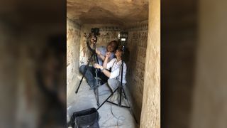 Two researchers crouch in an ancient Egyptian tomb that has wall paintings. They have a portable instrument that allows them to analyze the artwork.