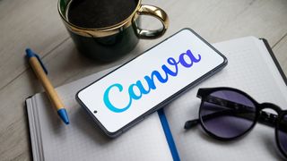 Canva logo and branding pictured on a smartphone sitting on a desk with notepad, coffee cup, and pen pictured alongside.