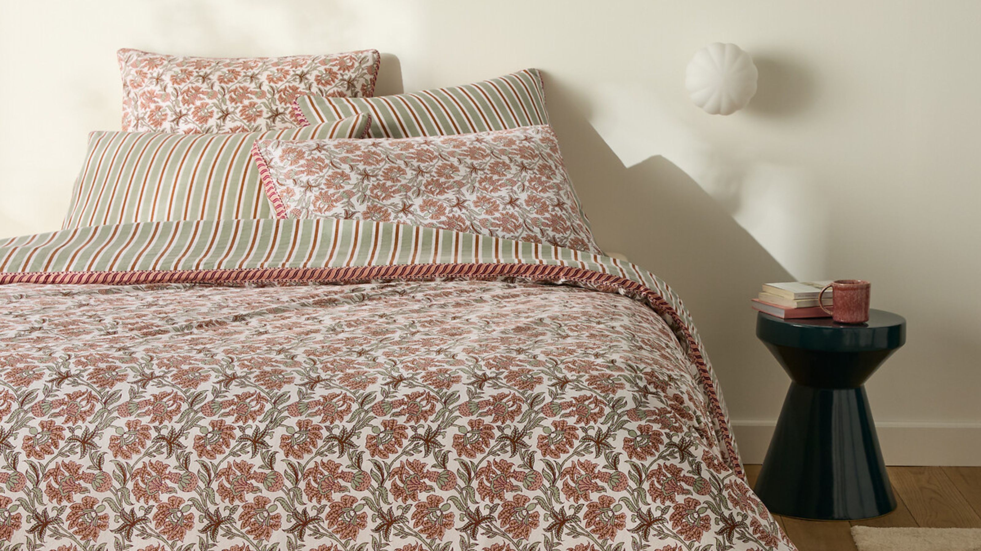 bed made with reversible bedding with floral print on one side and stripes on the other, with a black side table styled with books and a mug and a wall sconce above