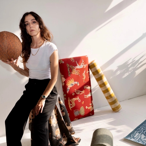 Image of a woman in a white top and black pants sitting in a white room with different rolls of patterned fabric and wallpaper.